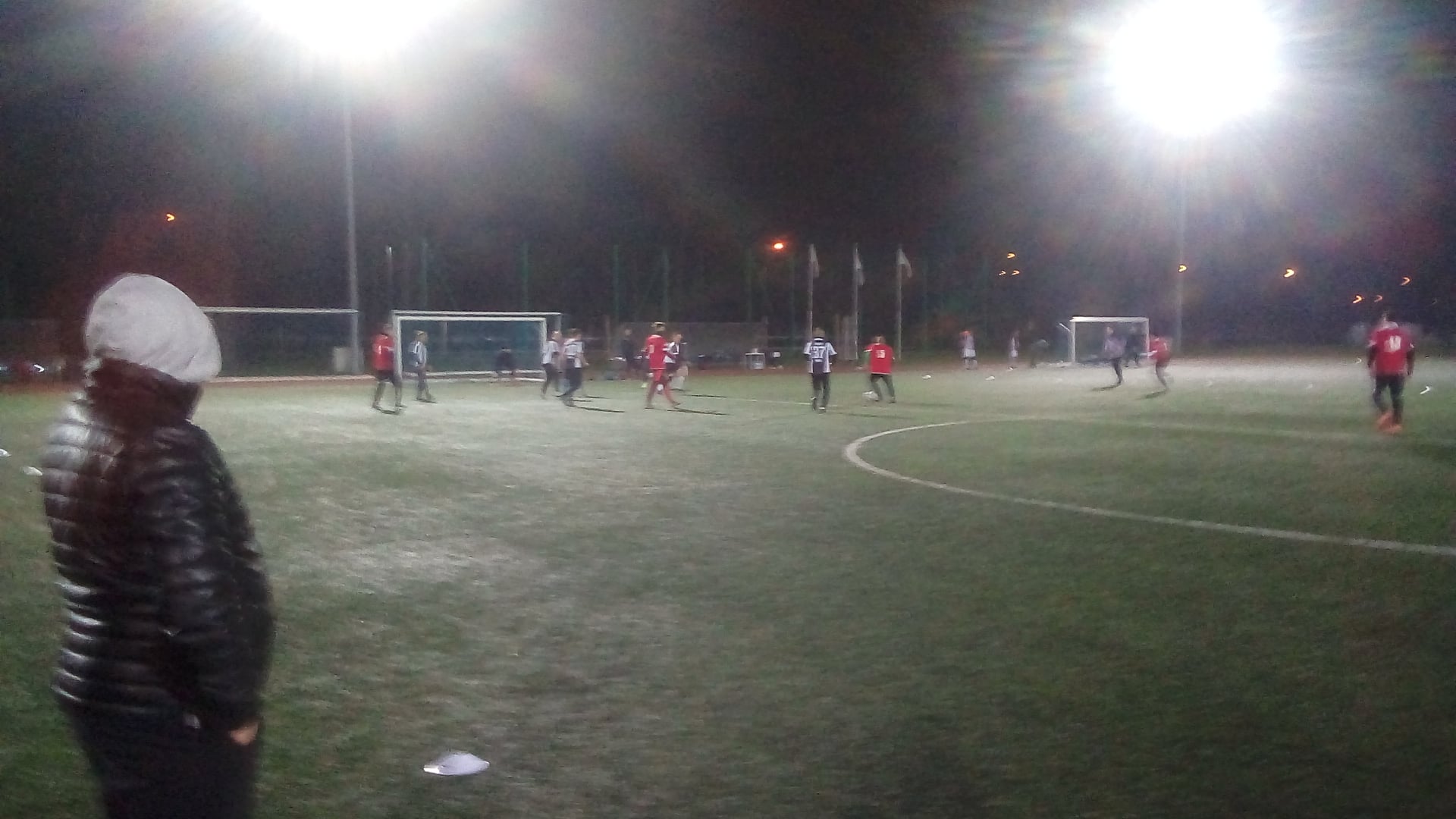 Piłkarze na zielonej murawie boiska piłkarskiego. Gra się toczy pod wieczór, w świetle reflektorów.
