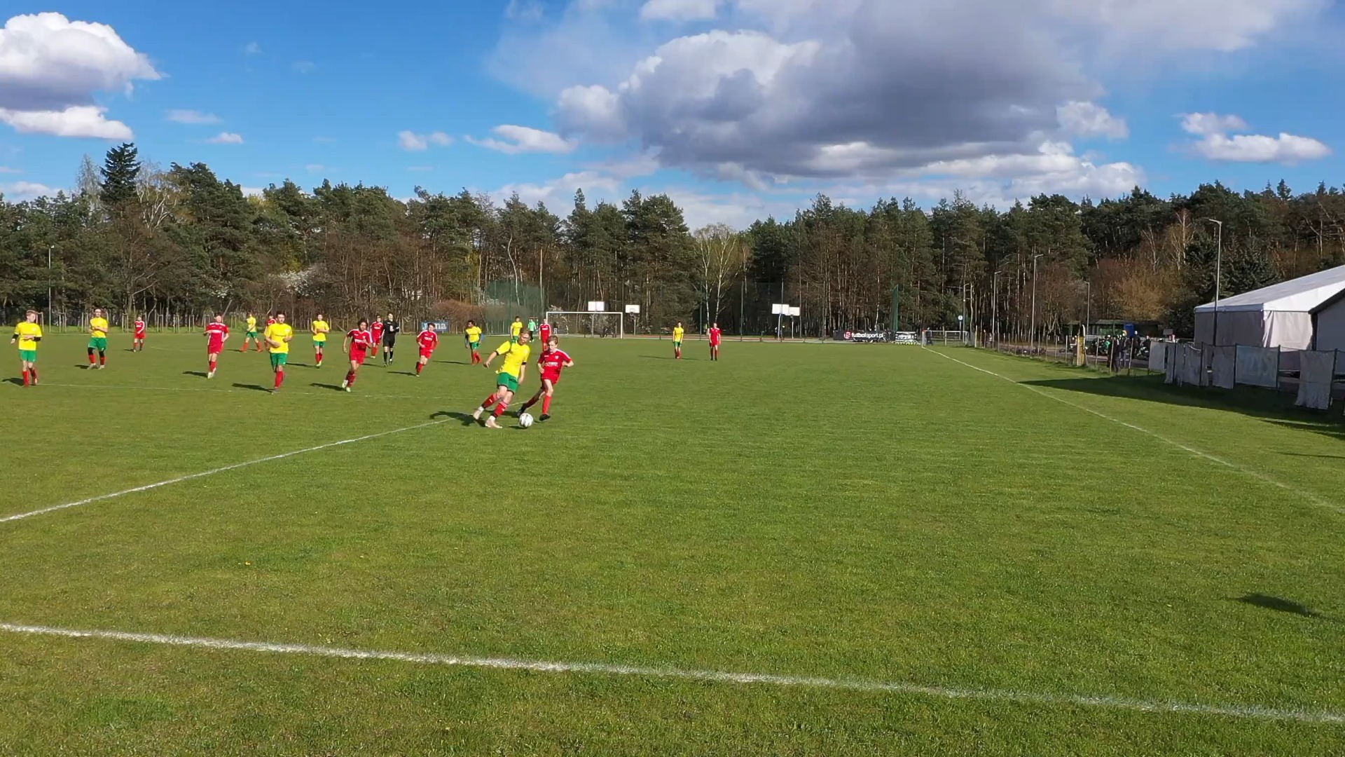 Piłkarze na zielonej murawie boiska piłkarskiego. Jedna z drużyn ma żółte stroje, druga czerwone.