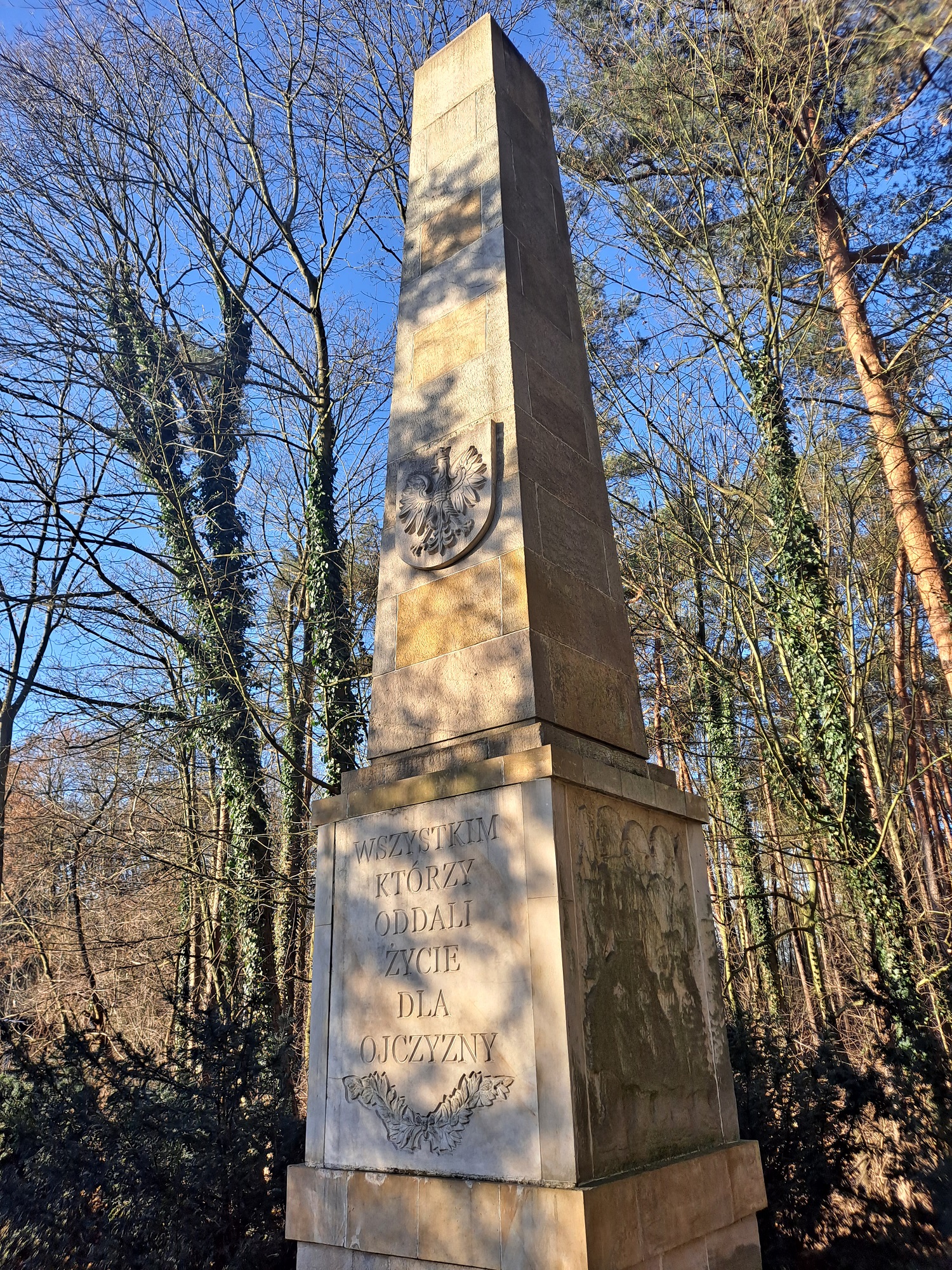 Obelisk pamiątkowy z napisem "Wszystkim, którzy oddali życie dla ojczyzny". Skąpany w słońcu, wśród drzew bez liści.